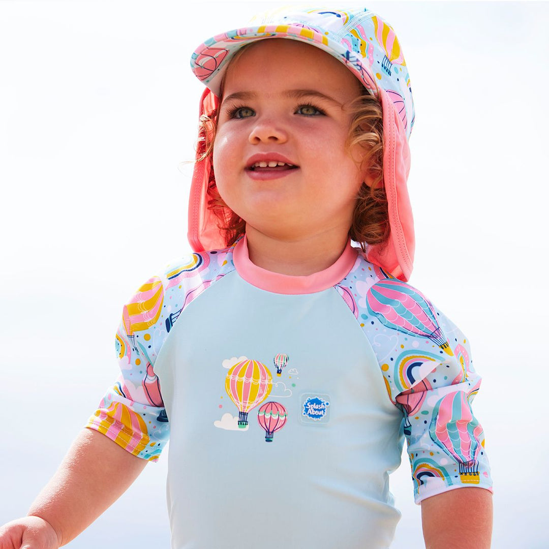 Lifestyle image of toddler wearing a legionnaire style sun hat in baby blue and pink, with hot air balloons themed print panel, including clouds and rainbows. She's also wearing a matching Happy Nappy Sunsuit.