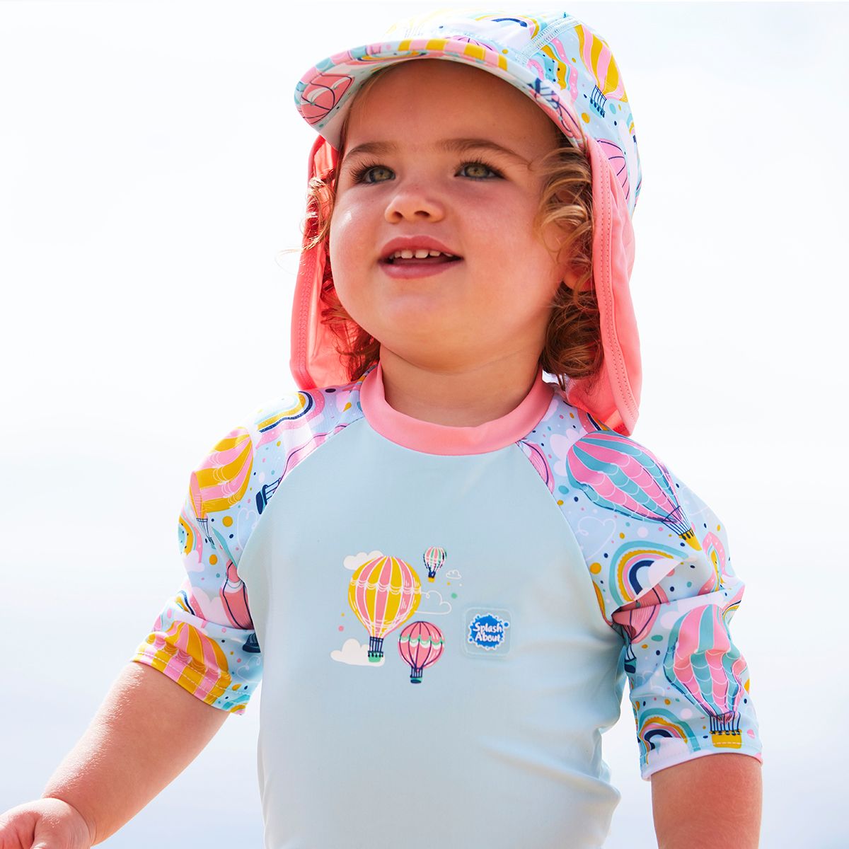 Lifestyle image of toddler wearing a legionnaire style sun hat in baby blue and pink, with hot air balloons themed print panel, including clouds and rainbows. She's also wearing a matching Happy Nappy Sunsuit.