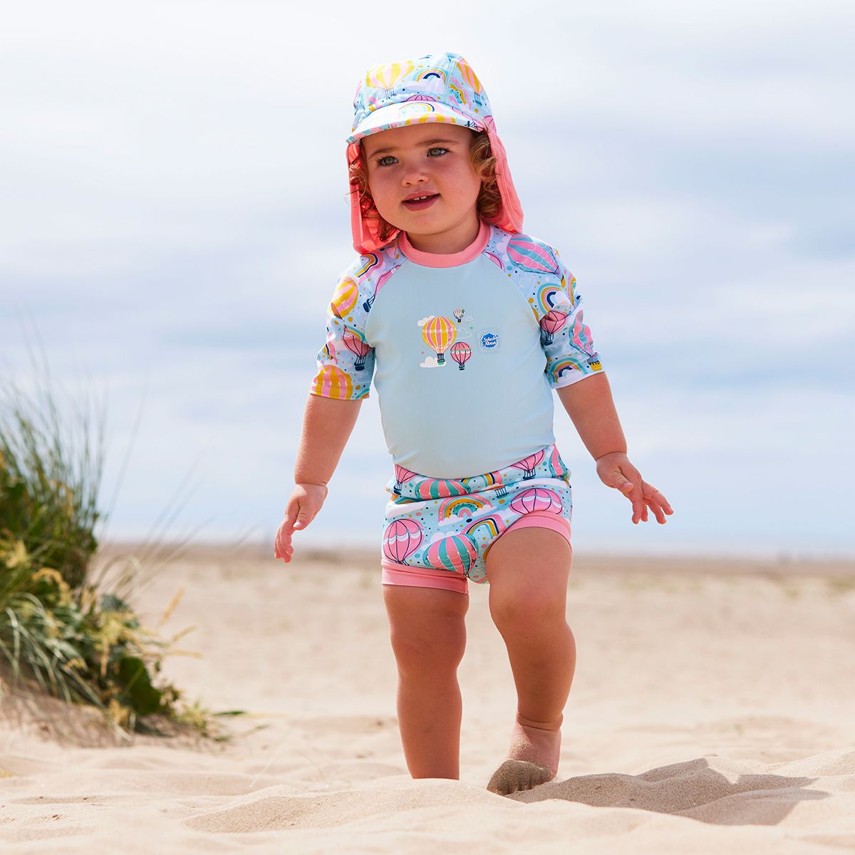 Lifestyle image of toddler wearing a legionnaire style sun hat in baby blue and pink, with hot air balloons themed print panel, including clouds and rainbows. She's also wearing a matching Happy Nappy Sunsuit.