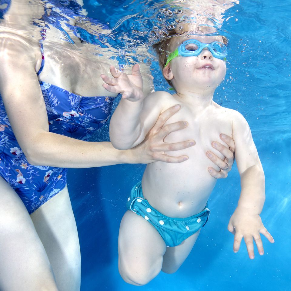 Lifestyle image of toddler wearing blue and green kids goggles with clear lenses. He's also wearing size adjustable blue under nappy.