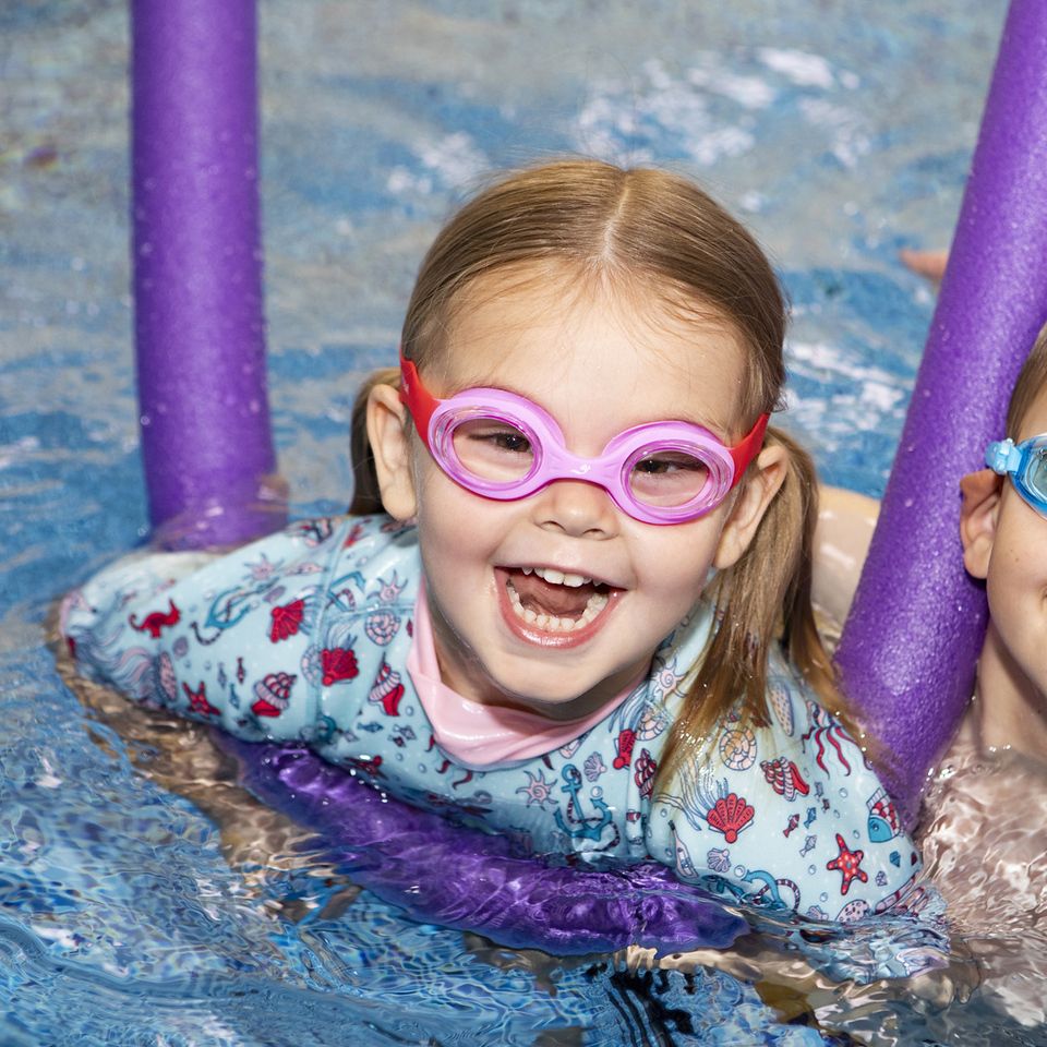 Lifestyle image of toddler wearing pink and red kids goggles with clear lenses. She's also wearing Hidden Treasure Happy Nappy wetsuit.