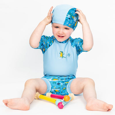 Toddler wearing  swim hat in blue with light blue trims and swamp themed print, including crocodiles, fireflies, frogs, snails and more. He's also wearing matching rash top and Happy Nappy.