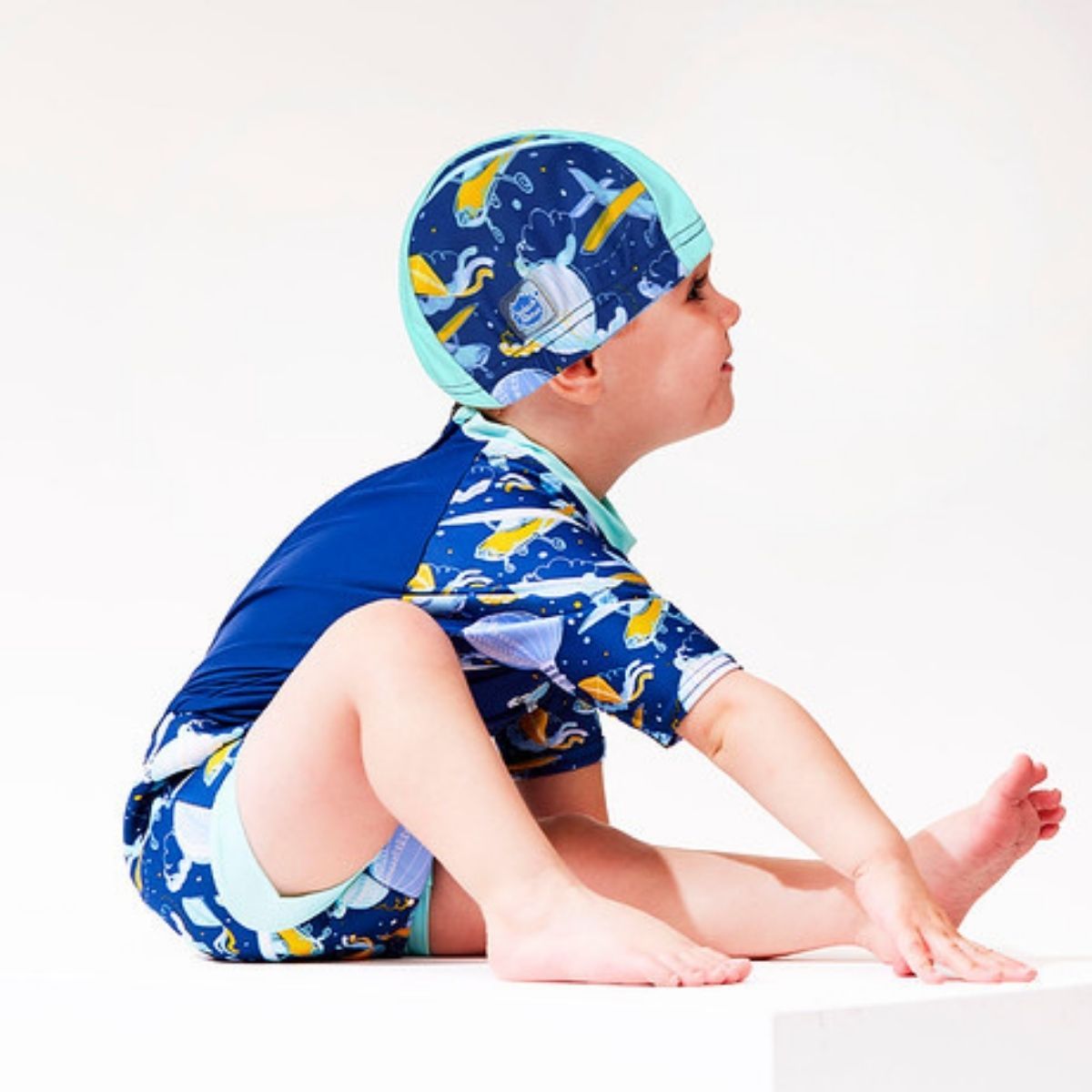 Toddler wearing  swim hat in blue with light green trims and sky themed print, including airplanes, kites, clouds, hot air balloons and more. He's also wearing matching rash top and Happy Nappy.