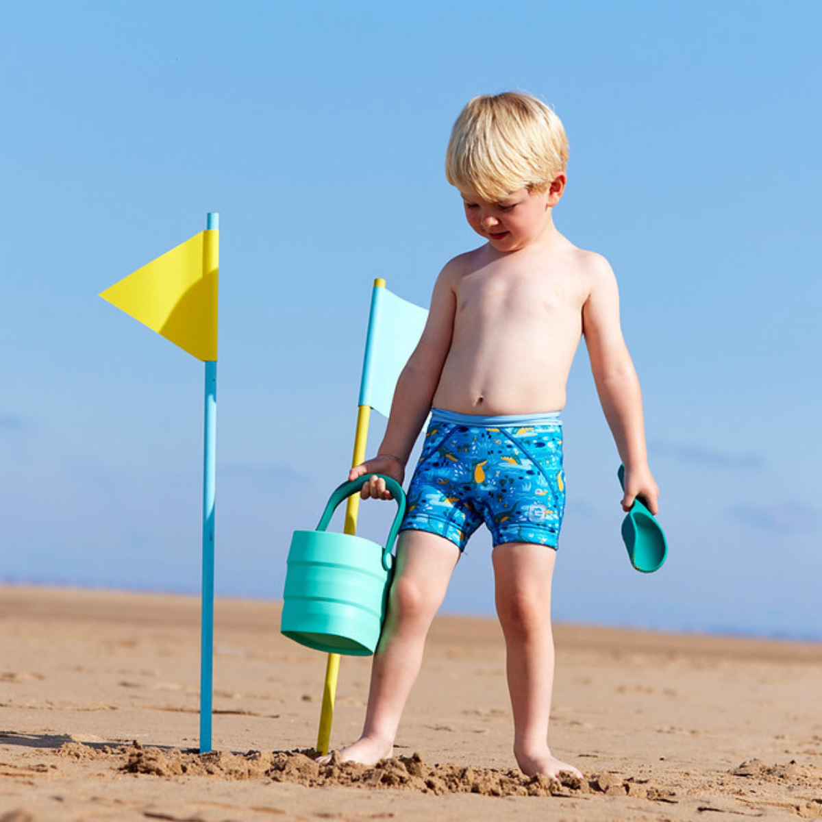 Lifestyle image of toddler sitting by the pool, wearing neoprene swim shorts in blue with light blue waist and swamp themed print, including crocodiles, frogs, snails and more.
