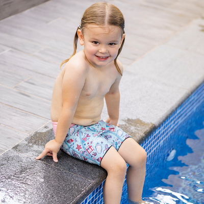 Lifestyle image of toddler sitting by the pool, wearing neoprene swim shorts in light blue with baby pink waist and sea themed print, including treasure chests, seahorses, fish, jellyfish and more.