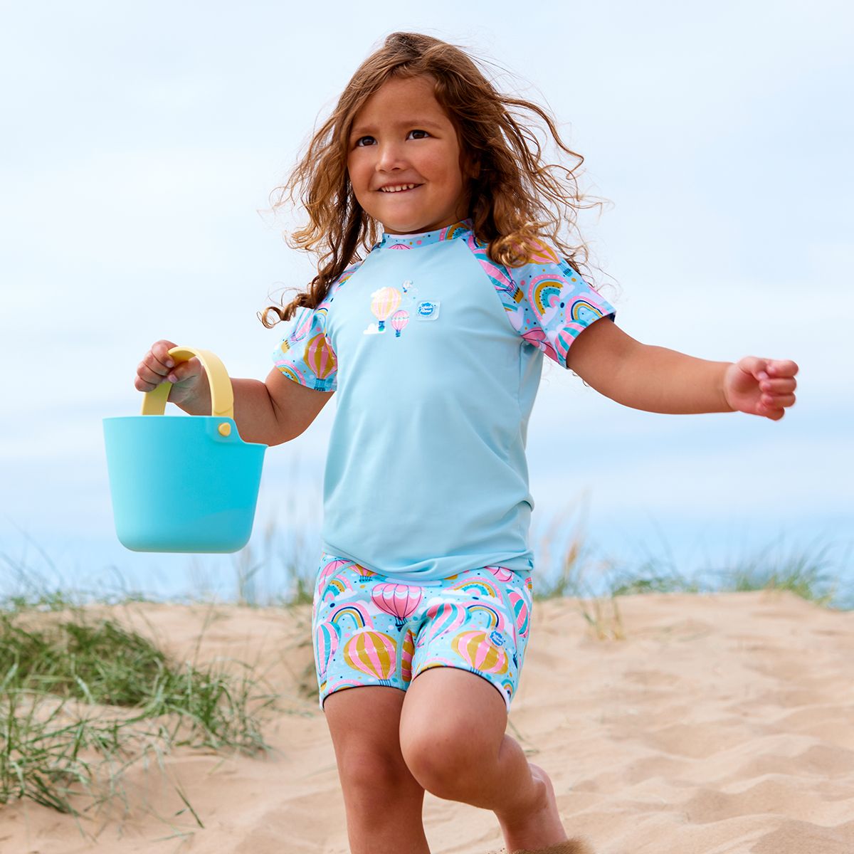 Lifestyle image of child wearing neoprene swim shorts in baby blue with pink waist and hot air balloons themed print, including clouds and rainbows. 