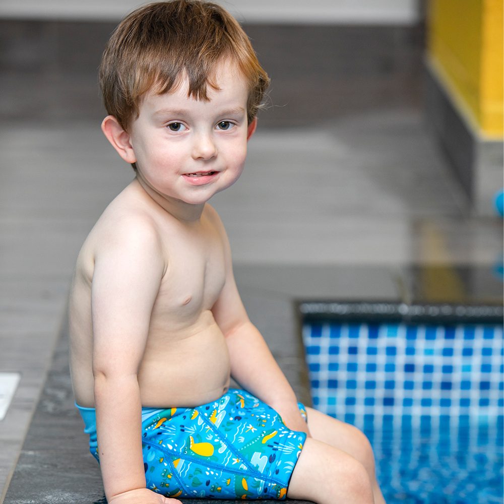 Lifestyle image of toddler sitting by the pool, wearing neoprene swim shorts in blue with light blue waist and swamp themed print, including crocodiles, frogs, snails and more.