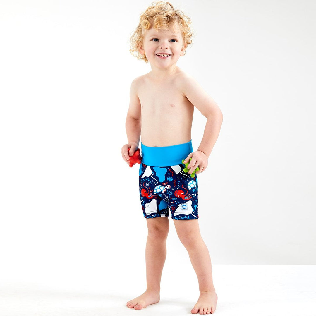 Lifestyle image of toddler wearing neoprene swim shorts in navy blue with blue waist and under the sea themed print, including octopus, turtles, fish, stingray and more.