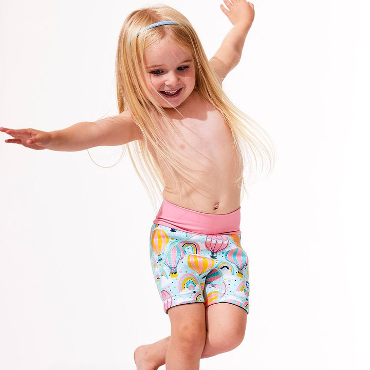 Lifestyle image of child wearing neoprene swim shorts in baby blue with pink waist and hot air balloons themed print, including clouds and rainbows. 