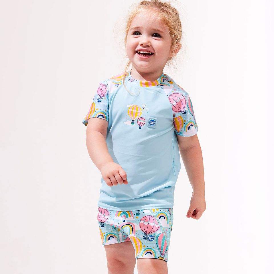Lifestyle image of toddler wearing UV protective short sleeve rash top in light blue, and hot air balloons print on the chest and sleeves. She's also wearing matching swim shorts jammers.
