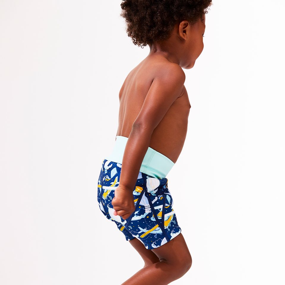 Lifestyle image of toddler wearing neoprene swim shorts in navy blue with light green waist and hot air balloons themed print, including airplanes, kites and clouds.