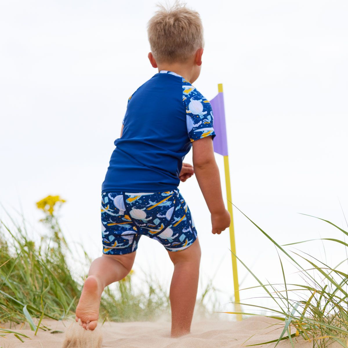 Lifestyle image of toddler running in the beach, wearing neoprene swim shorts in navy blue with light green waist and hot air balloons themed print, including airplanes, kites and clouds. Back of the jammers, he's also wearing matching rash top.