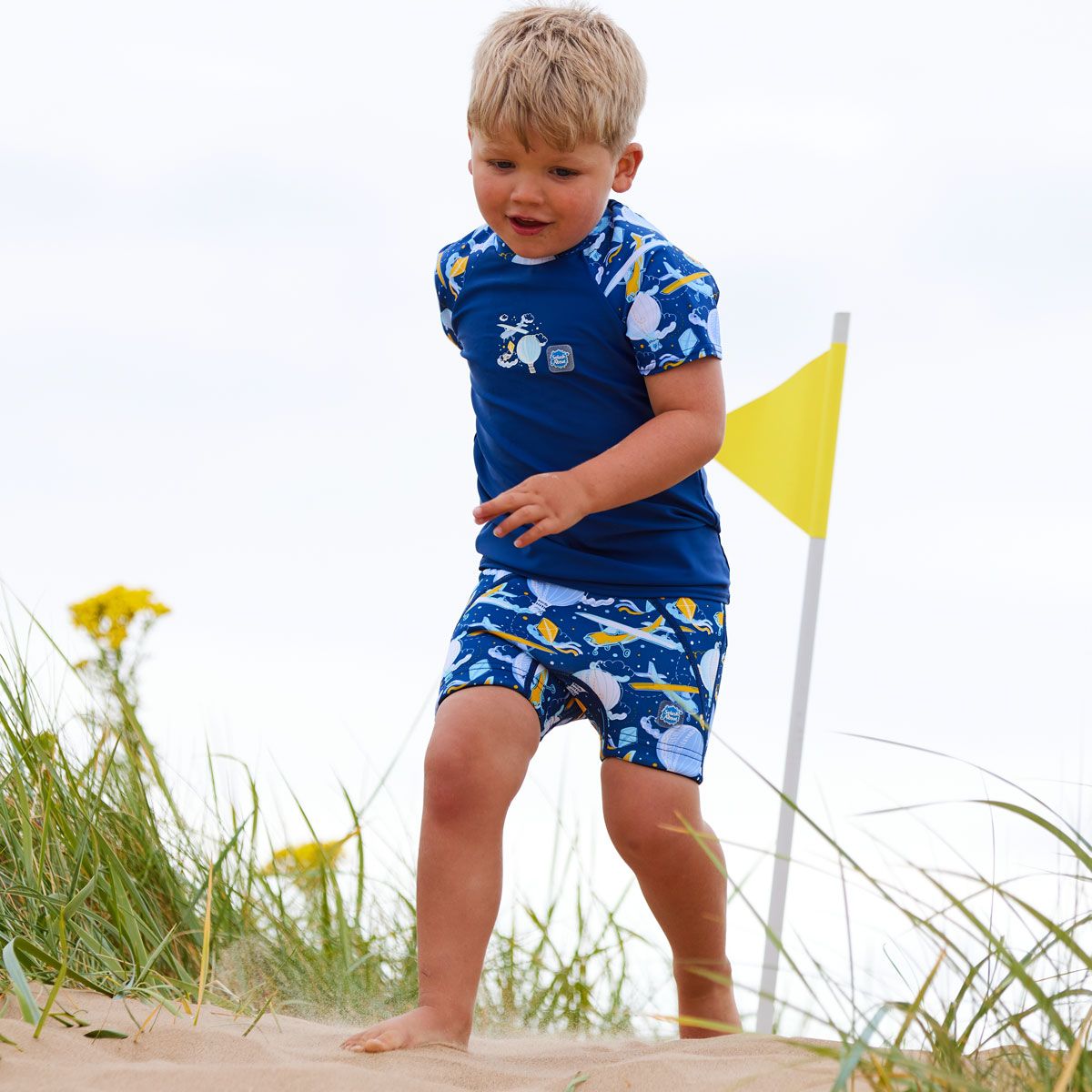 Lifestyle image of toddler running in the beach, wearing neoprene swim shorts in navy blue with light green waist and hot air balloons themed print, including airplanes, kites and clouds. Front of the jammers, he's also wearing matching rash top.