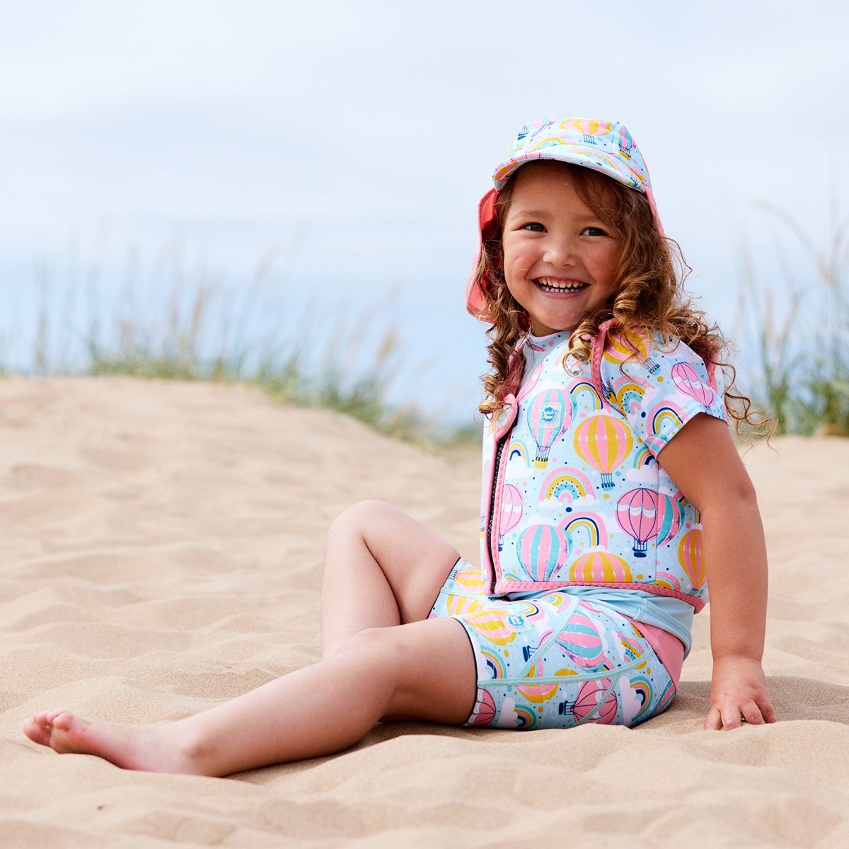 Lifestyle image of child wearing neoprene swim vest for toddlers with non-removable floats in baby blue, pink trims and hot air balloons themed print, including clouds and rainbows. She's also wearing matching legionnaire hat, jammers and rash top.