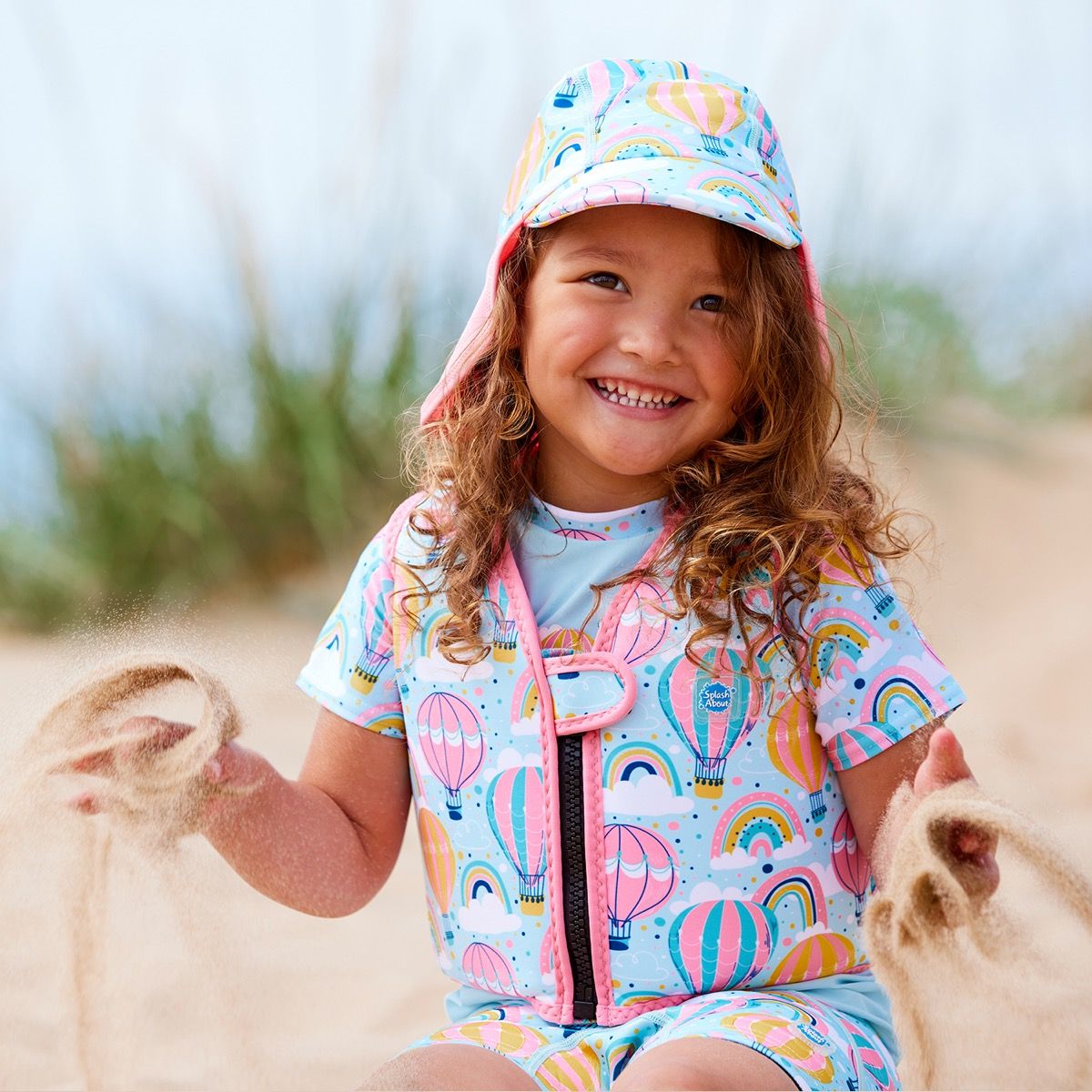 Lifestyle image of child wearing neoprene swim vest for toddlers with non-removable floats in baby blue, pink trims and hot air balloons themed print, including clouds and rainbows. She's also wearing matching legionnaire hat, jammers and rash top.