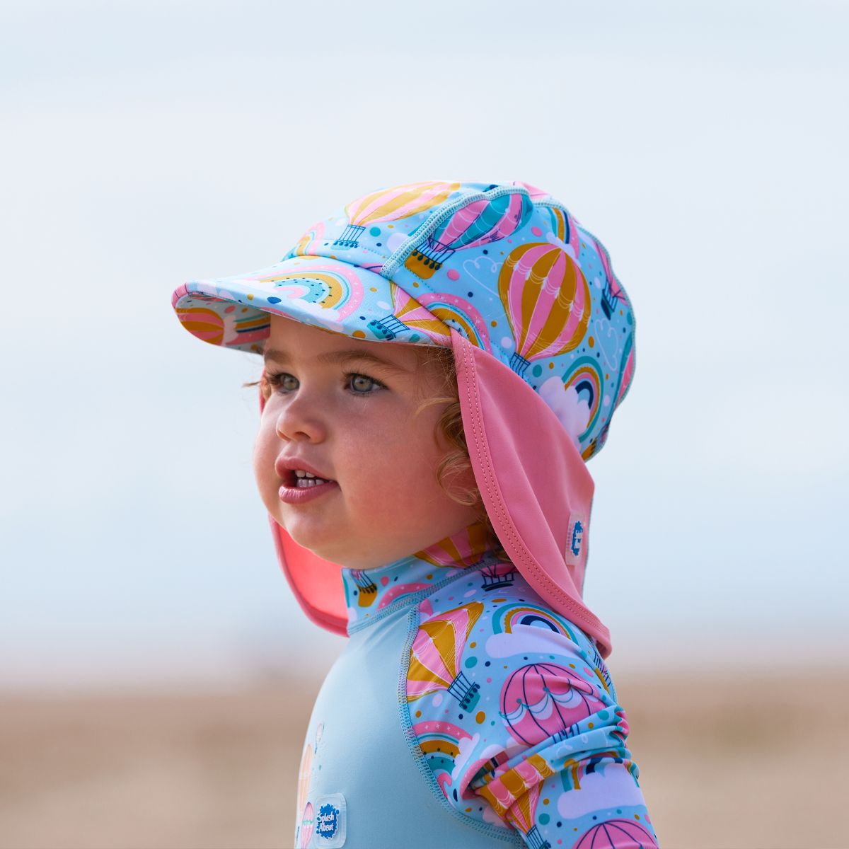 Lifestyle image of toddler wearing a legionnaire style sun hat in baby blue and pink, with hot air balloons themed print panel, including clouds and rainbows. She's also wearing a matching Sun & Sea Wetsuit.