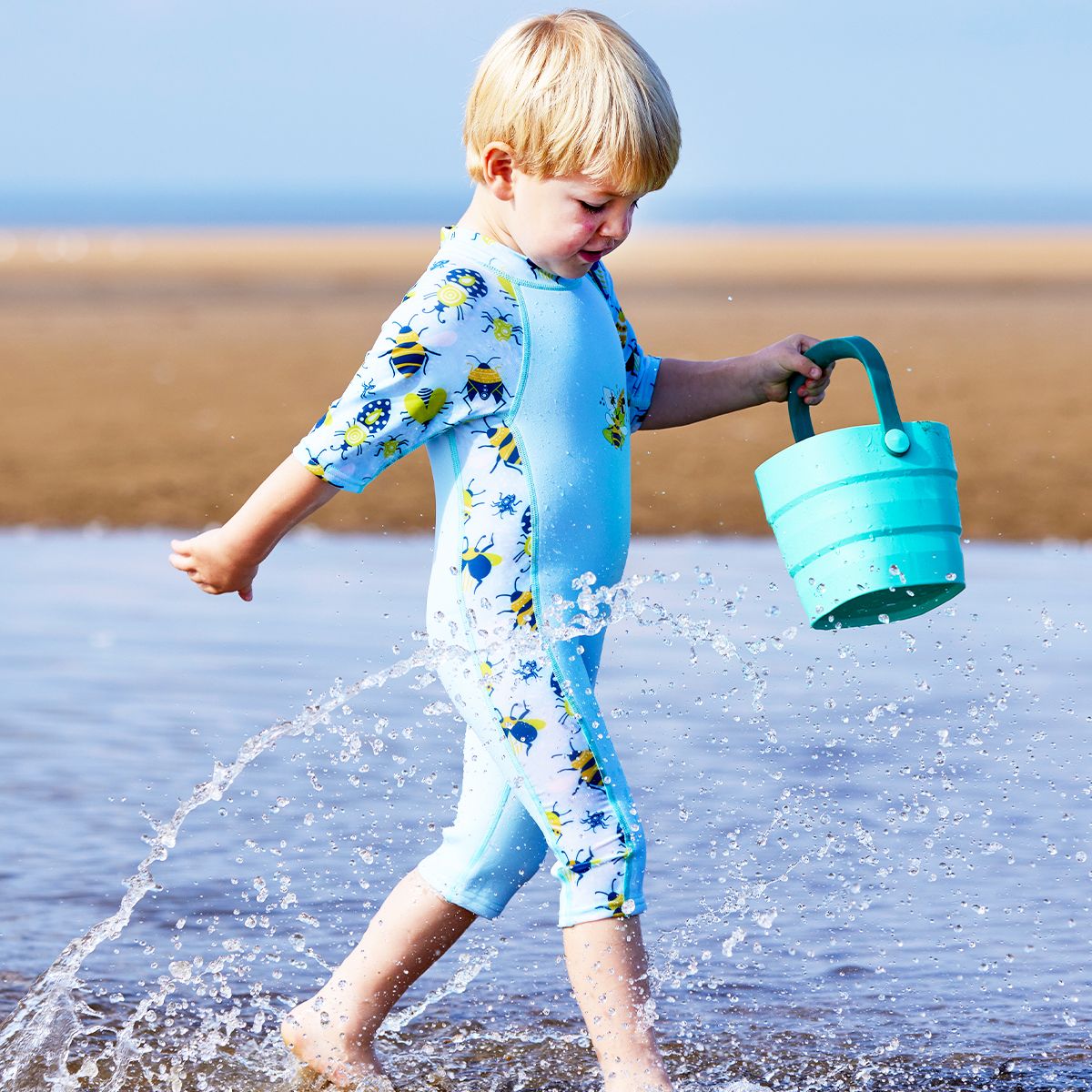 Lifestyle image of child wearing a one piece UV sun and sea wetsuit for toddlers in light blue with navy blue trims. Insects themed print on sleeves, side panels and neck. 