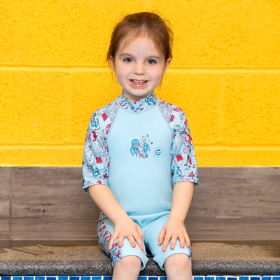 Lifestyle image of child wearing a one piece UV sun and sea wetsuit for toddlers in light blue and sea life themed print including starfish, fish, anchors, jellyfish, seahorses and more on sleeves, side panels and neck.