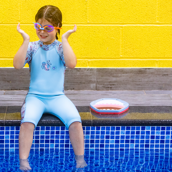Lifestyle image of child wearing a one piece UV sun and sea wetsuit for toddlers in light blue and sea life themed print including starfish, fish, anchors, jellyfish, seahorses and more on sleeves, side panels and neck.