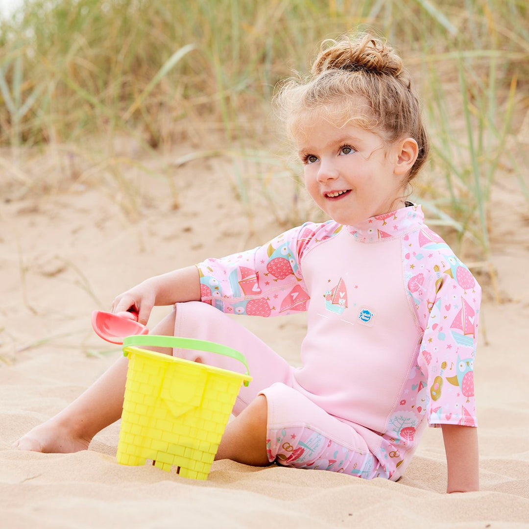 Lifestyle image of child wearing a one piece UV sun and sea wetsuit for toddlers in baby pink. Cute kittens, owls, trees, guitars and seal boats print on sleeves, side panels and neck. 