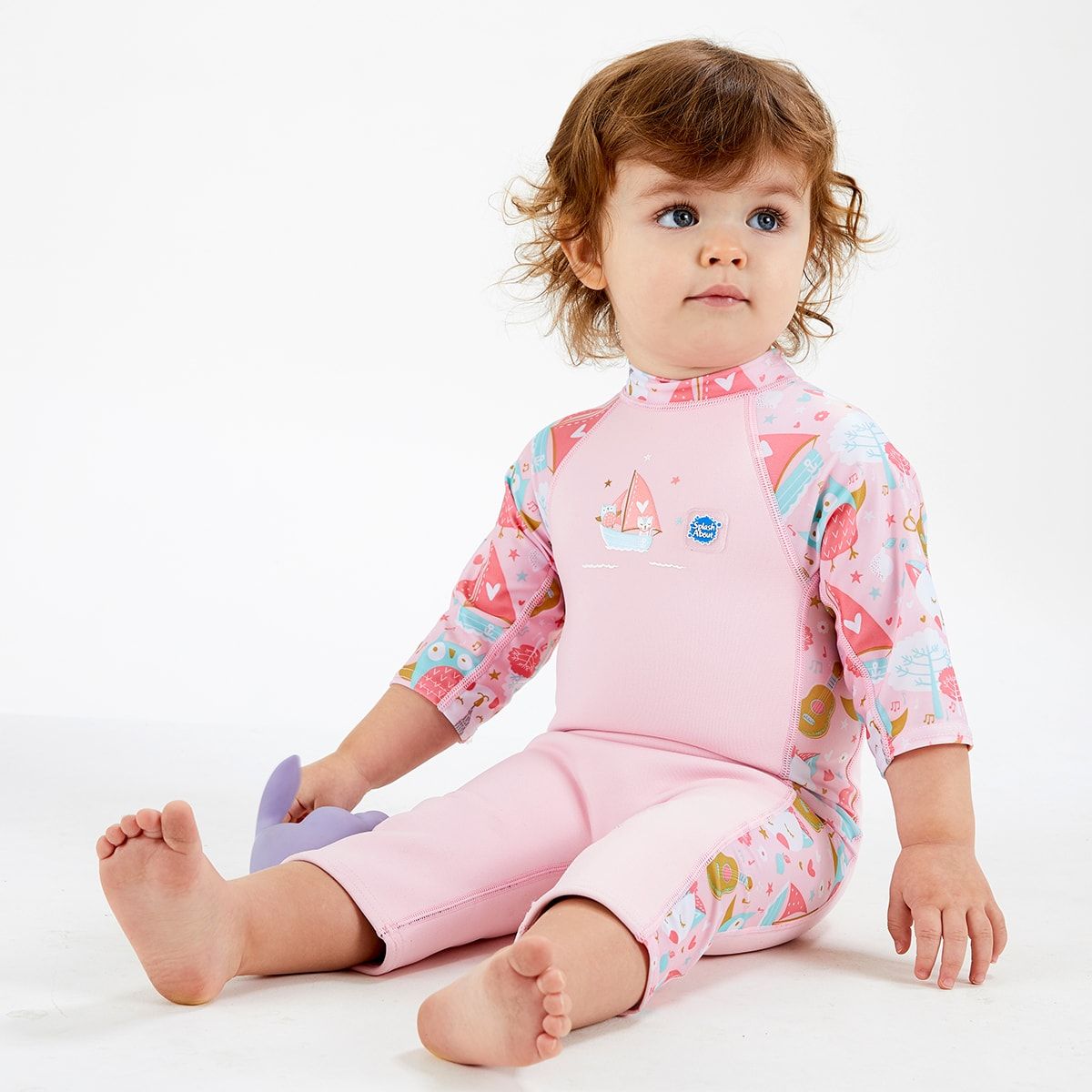 Lifestyle image of child wearing a one piece UV sun and sea wetsuit for toddlers in baby pink. Cute kittens, owls, trees, guitars and seal boats print on sleeves, side panels and neck. 