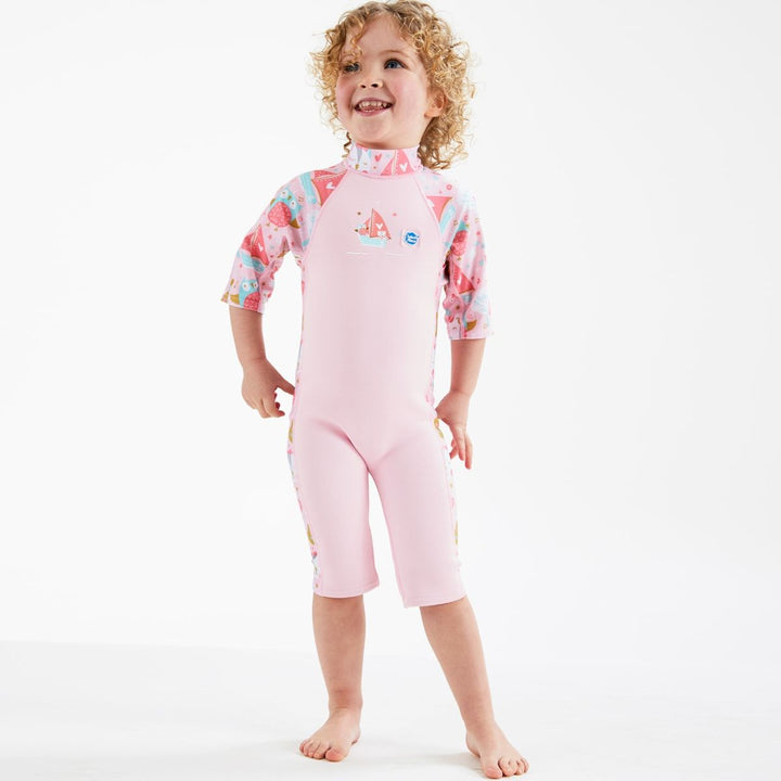 Lifestyle image of child wearing a one piece UV sun and sea wetsuit for toddlers in baby pink. Cute kittens, owls, trees, guitars and seal boats print on sleeves, side panels and neck. 