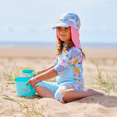 Lifestyle image of a child wearing a one piece UV sun and sea wetsuit for toddlers in light blue. Hot air balloons themed print including clouds and rainbows on sleeves, side panels, neck and chest. She's also wearing a matching legionnaire hat.