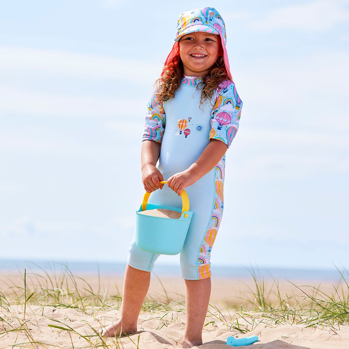 Lifestyle image of a child wearing a one piece UV sun and sea wetsuit for toddlers in light blue. Hot air balloons themed print including clouds and rainbows on sleeves, side panels, neck and chest. She's also wearing a matching legionnaire hat.