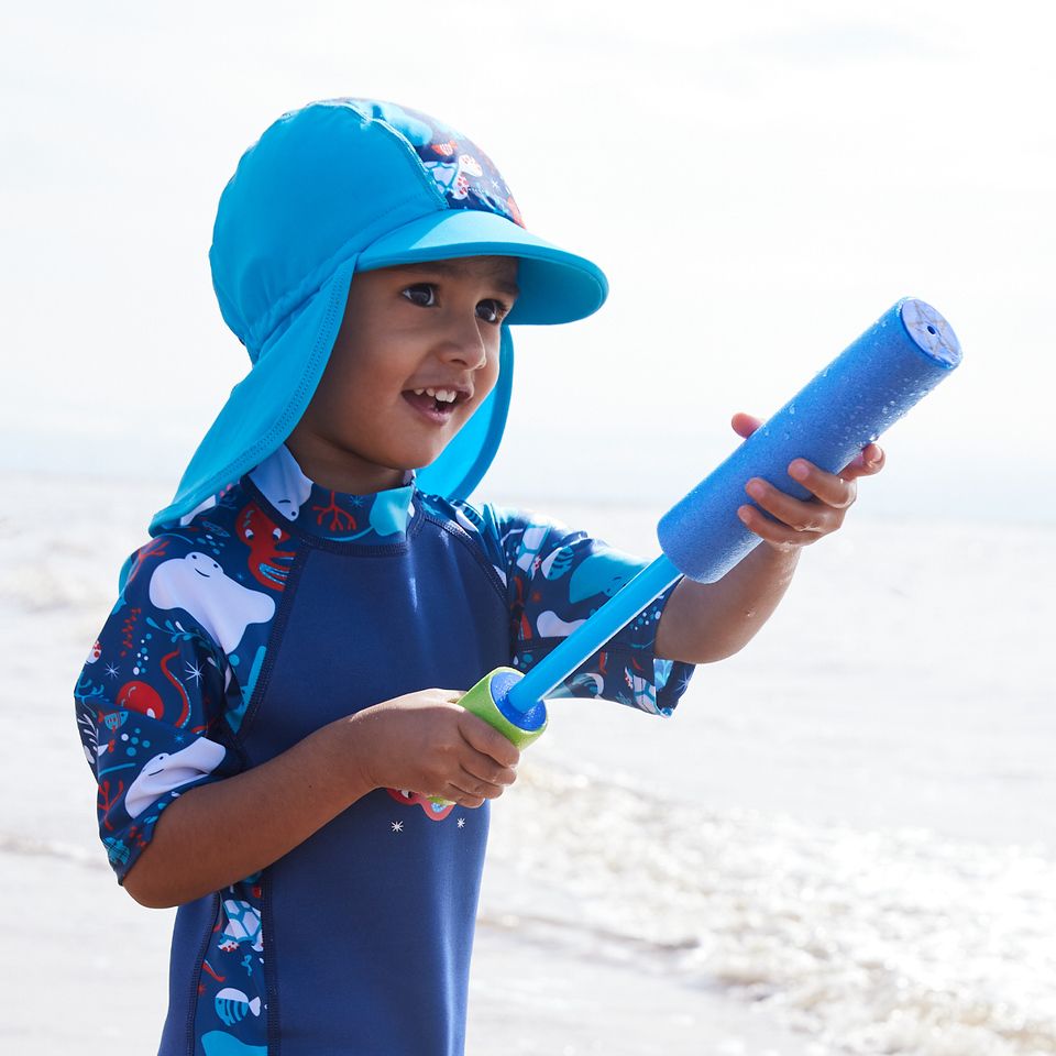 Lifestyle image of child wearing a one piece UV sun and sea wetsuit for toddlers in navy blue with cyan trims. Sea life themed print including turtles, octopus, fish, stingray and more on sleeves, side panels and neck. He's also wearing a matching legionnaire hat.