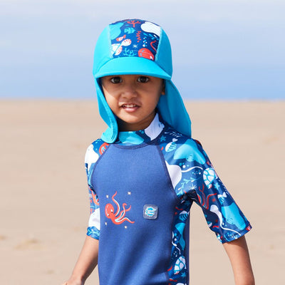 Lifestyle image of child wearing a one piece UV sun and sea wetsuit for toddlers in navy blue with cyan trims. Sea life themed print including turtles, octopus, fish, stingray and more on sleeves, side panels and neck. He's also wearing a matching legionnaire hat.