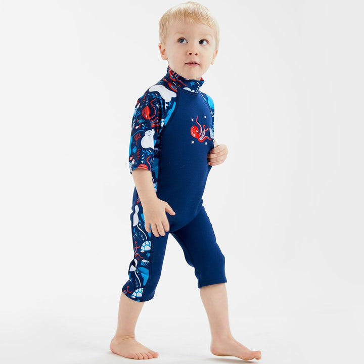 Lifestyle image of child wearing a one piece UV sun and sea wetsuit for toddlers in navy blue with cyan trims. Sea life themed print including turtles, octopus, fish, stingray and more on sleeves, side panels and neck.