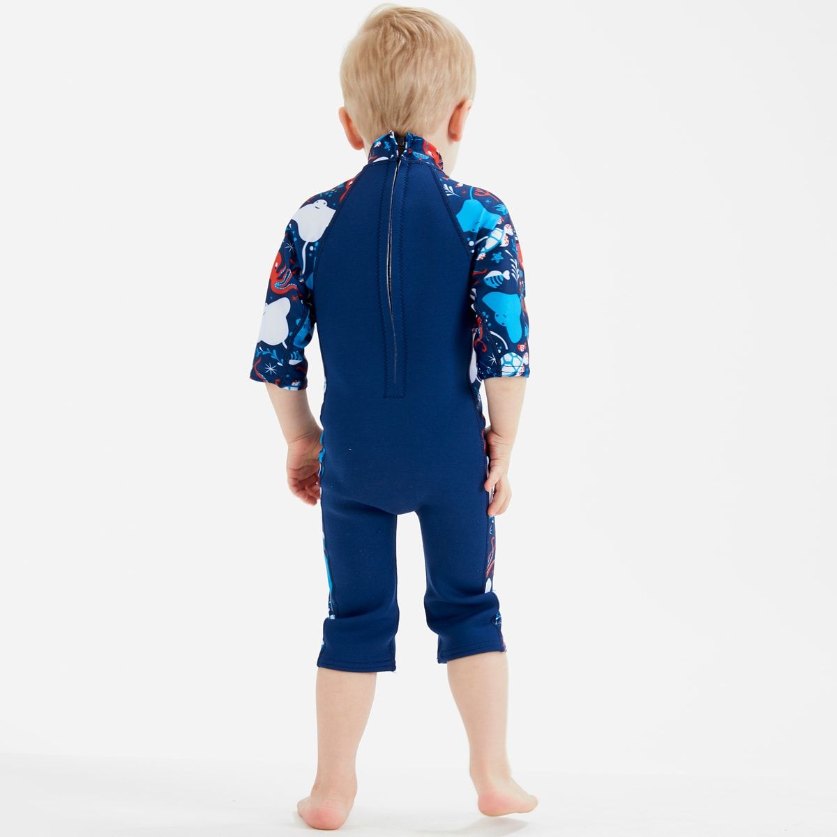 Lifestyle image of child wearing a one piece UV sun and sea wetsuit for toddlers in navy blue with cyan trims. Sea life themed print including turtles, octopus, fish, stingray and more on sleeves, side panels and neck.