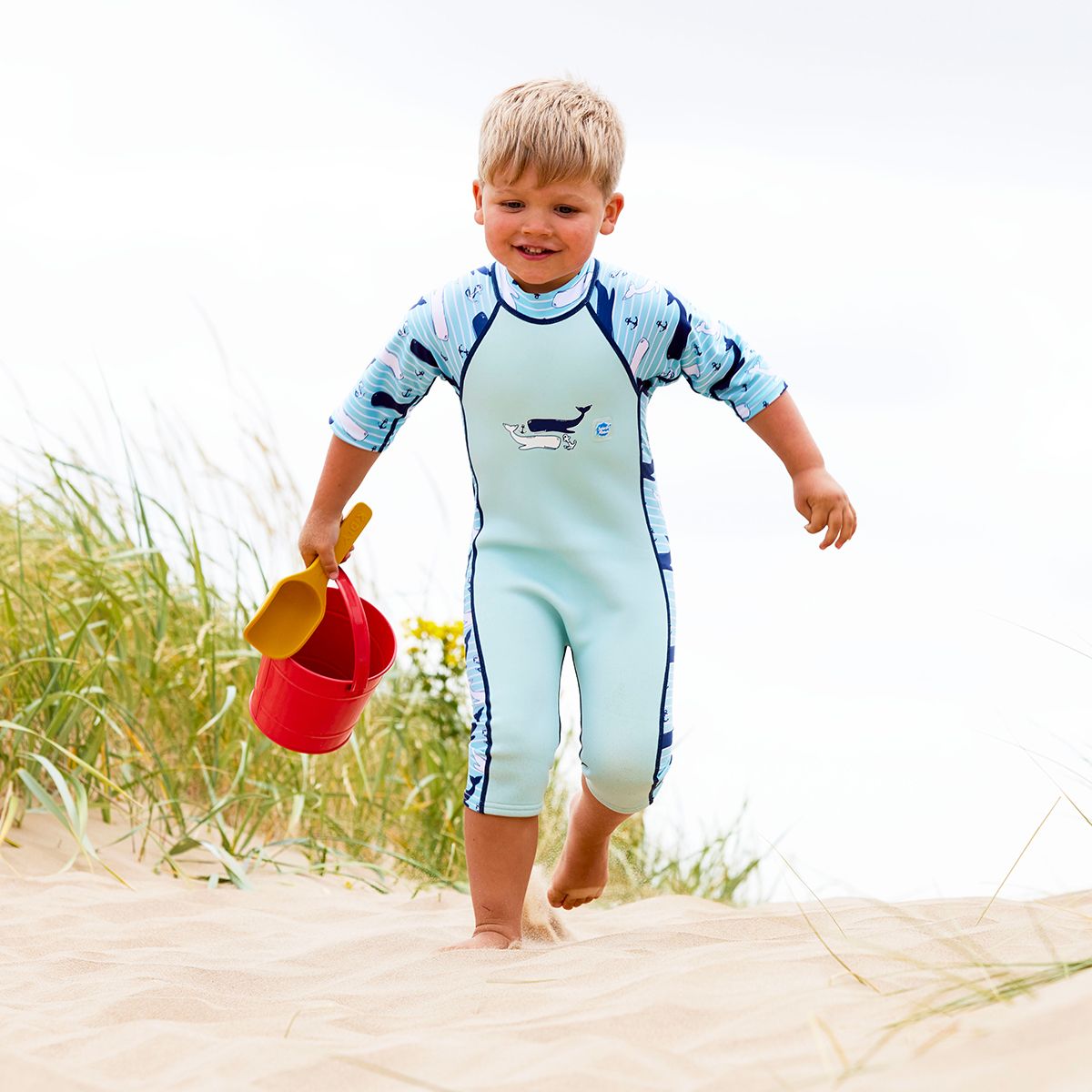Lifestyle image of child wearing a one piece UV sun and sea wetsuit for toddlers in light blue with navy blue trims. Stripes, whales and anchors print on sleeves, side panels and neck.
