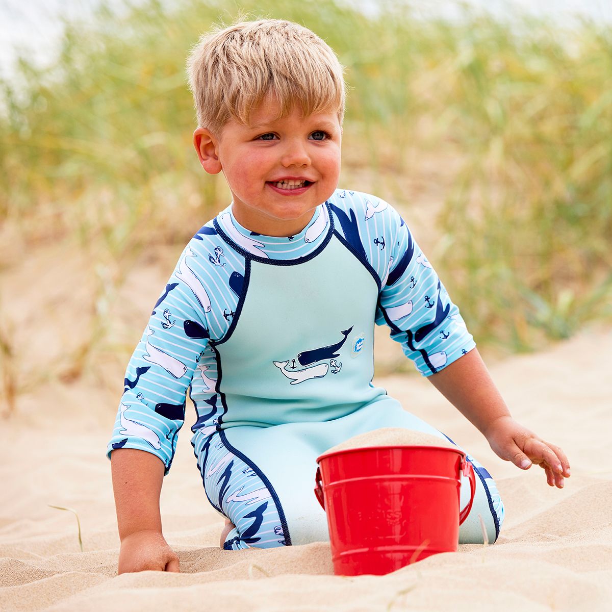 Lifestyle image of child wearing a one piece UV sun and sea wetsuit for toddlers in light blue with navy blue trims. Stripes, whales and anchors print on sleeves, side panels and neck.