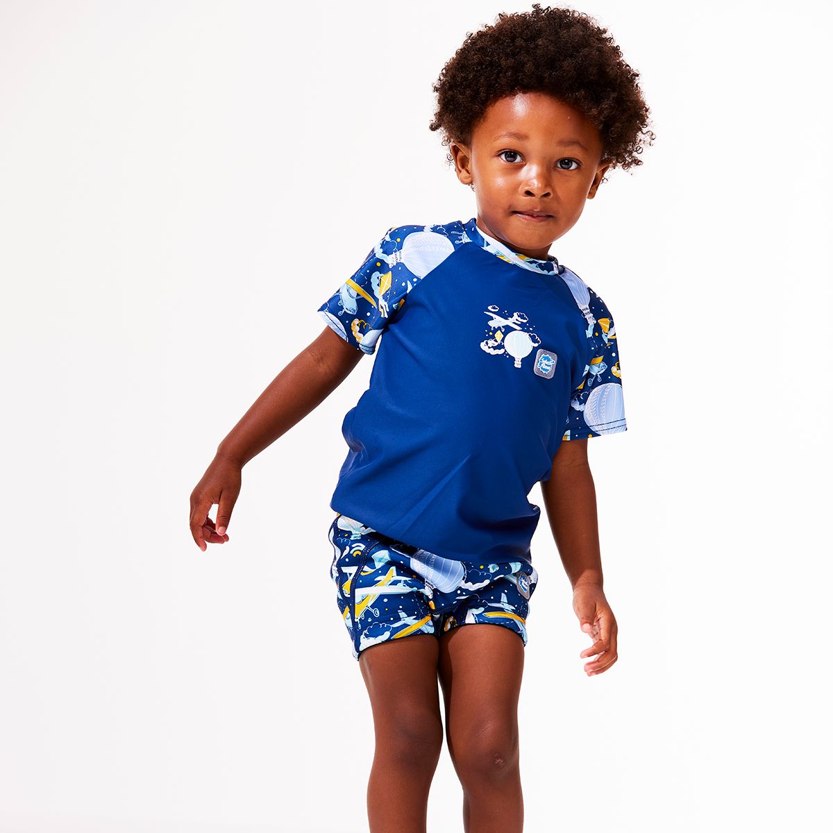 Lifestyle image of toddler wearing UV protective short sleeve rash top in navy blue, and sky themed print on the chest and sleeves. He's also wearing matching swim shorts or jammers.