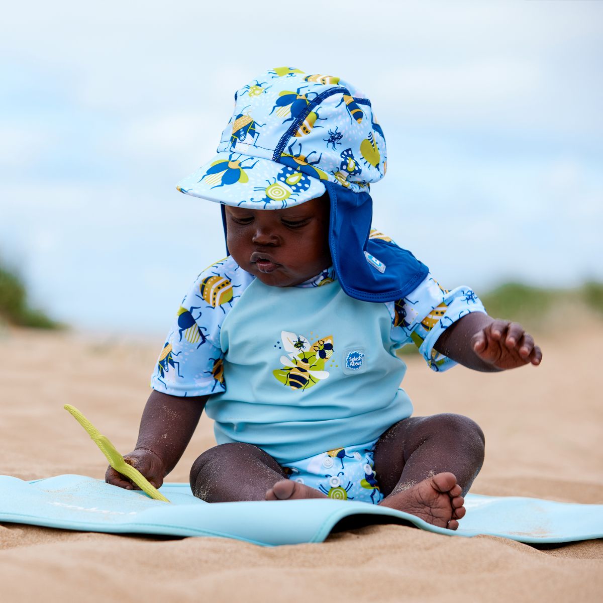 Lifestyle image of toddler wearing a legionnaire style sun hat in the beach. The hat is light blue and navy blue, with insects themed print panel. He's also wearing matching Happy Nappy and rash top.