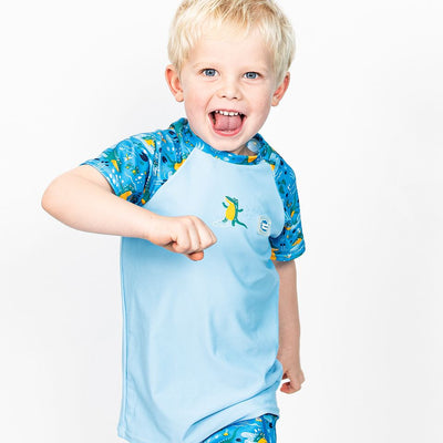 Lifestyle image of toddler wearing UV protective short sleeve rash top in light blue, and crocodile swamp themed print on the chest and sleeves, including crocodiles, frogs, fireflies. He's also wearing matching jammers.