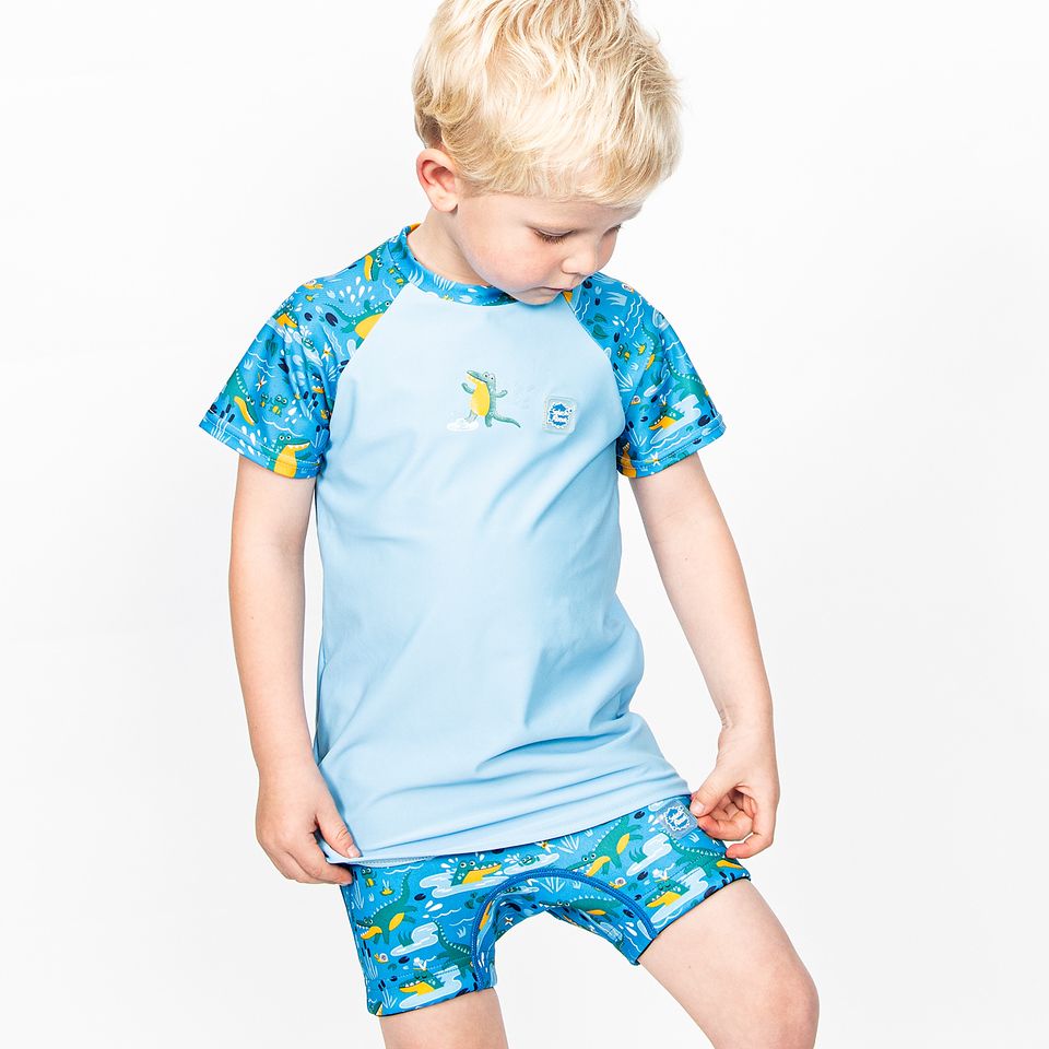 Lifestyle image of toddler wearing UV protective short sleeve rash top in navy blue, and crocodile swamp themed print on the chest and sleeves, including crocodiles, frogs, fireflies. He's also wearing matching jammers.