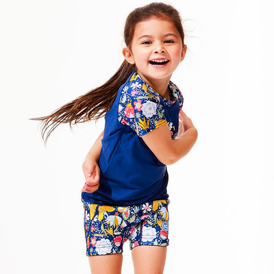 Lifestyle image of toddler wearing UV protective short sleeve rash top in navy blue, and floral print on the chest and sleeves. She's also wearing matching jammers.