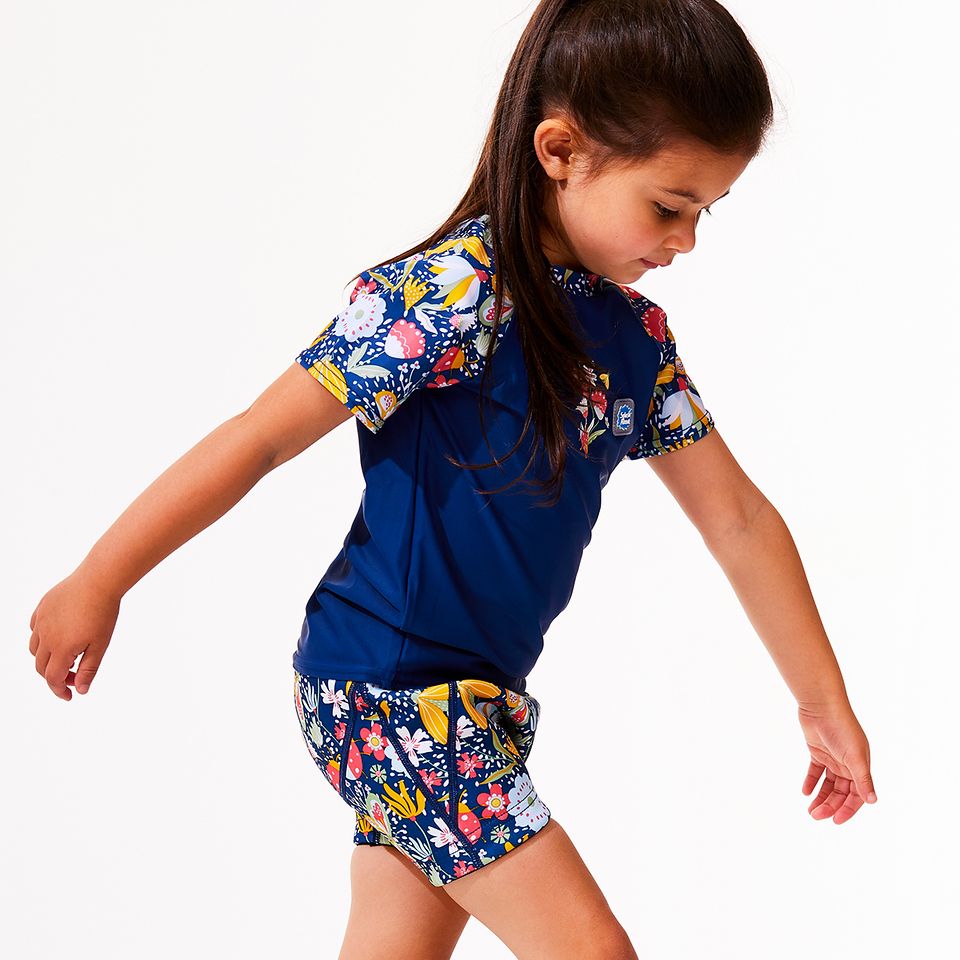 Lifestyle image of toddler wearing UV protective short sleeve rash top in navy blue, and floral print on the chest and sleeves. She's also wearing matching swim shorts or jammers.