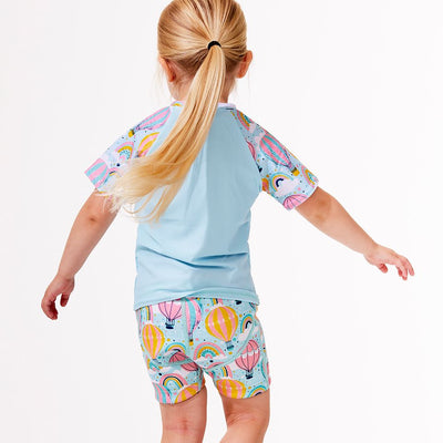Lifestyle image of toddler wearing UV protective short sleeve rash top in light blue, and hot air balloons print on the chest and sleeves. She's also wearing matching swim shorts jammers. Back.