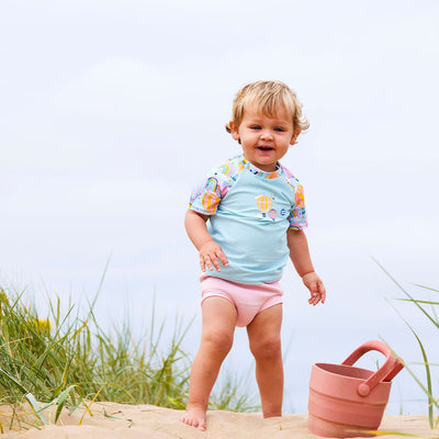 Lifestyle image of toddler wearing UV protective short sleeve rash top in light blue, and hot air balloons print on the chest and sleeves. He's also wearing the Almond Blossom Happy Nappy.