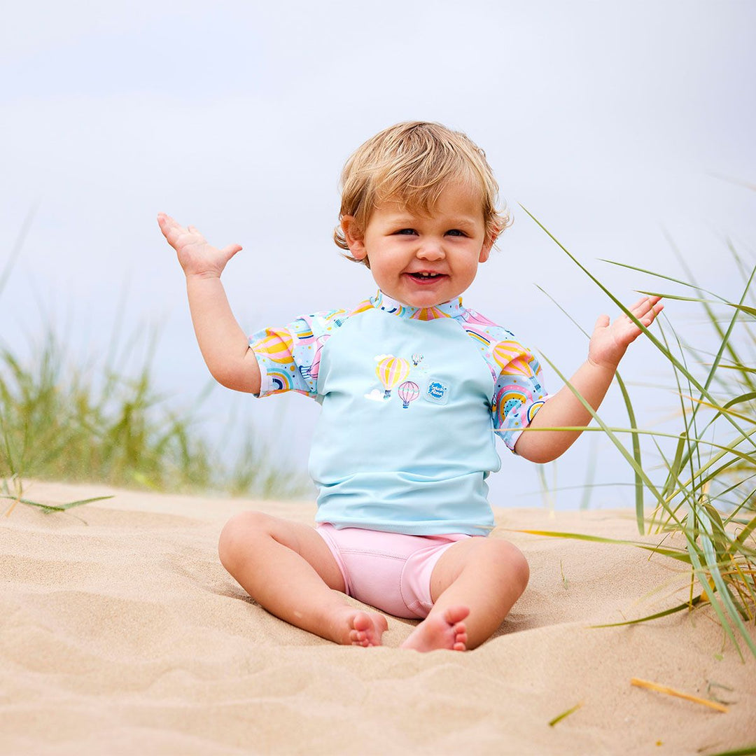 Lifestyle image of toddler wearing UV protective short sleeve rash top in light blue, and hot air balloons print on the chest and sleeves. He's also wearing the Almond Blossom Happy Nappy.