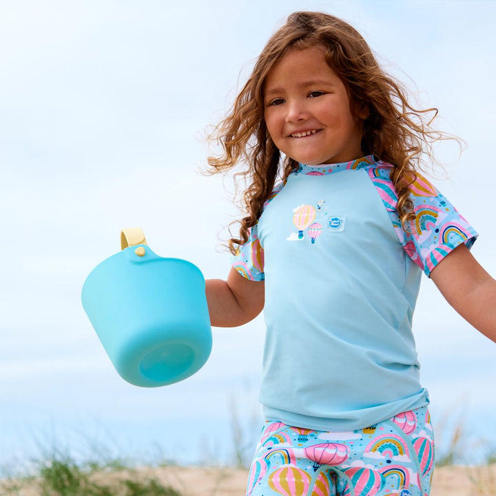 Lifestyle image of toddler wearing UV protective short sleeve rash top in light blue, and hot air balloons print on the chest and sleeves. She's also wearing matching jammers.