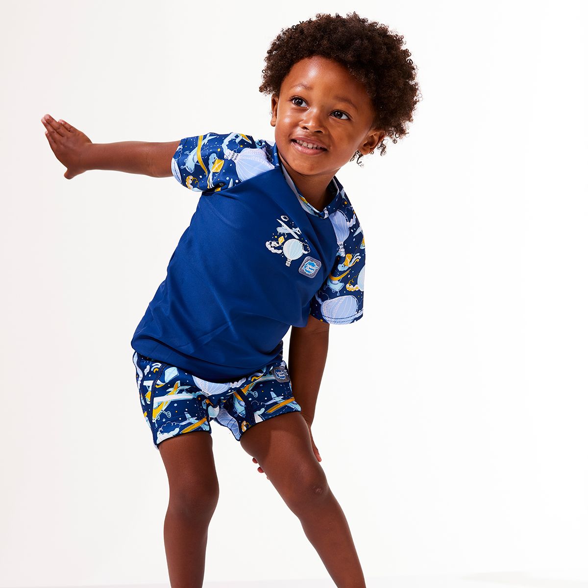 Lifestyle image of toddler wearing UV protective short sleeve rash top in navy blue, and sky themed print on the chest and sleeves. He's also wearing matching swim shorts or jammers.