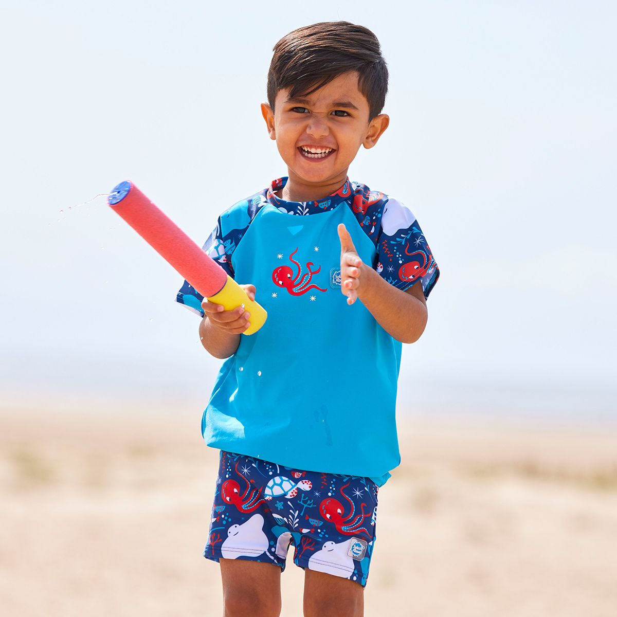 Lifestyle image of toddler wearing UV protective short sleeve rash top in blue, and under the sea themed print on the chest and sleeves. He's also wearing matching jammers or swim shorts.