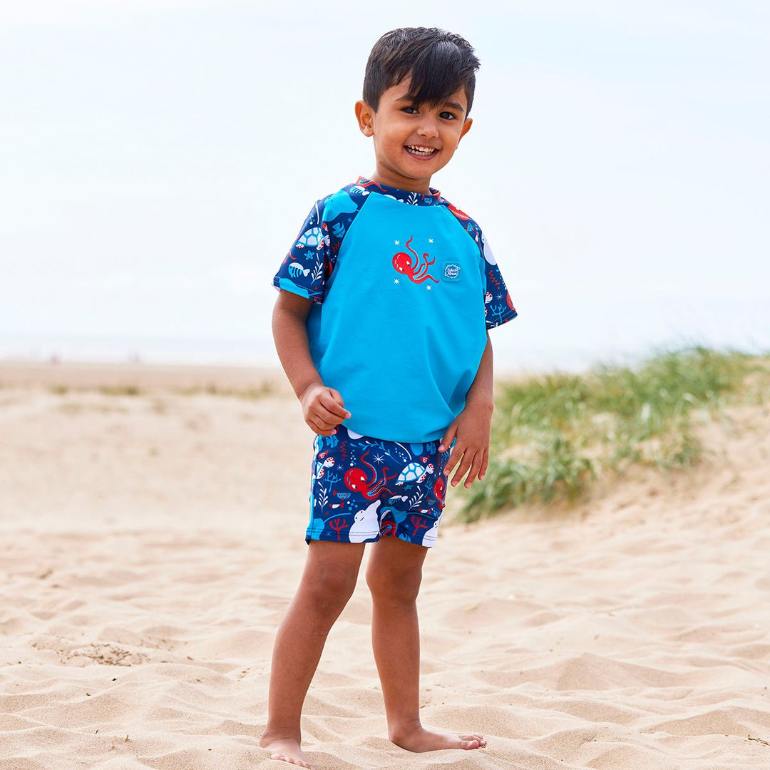 Lifestyle image of toddler wearing UV protective short sleeve rash top in blue, and under the sea themed print on the chest and sleeves. He's also wearing matching jammers or swim shorts.