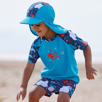 Lifestyle image of toddler wearing UV protective short sleeve rash top in blue, and under the sea themed print on the chest and sleeves. He's also wearing matching jammers and legionnaire hat.