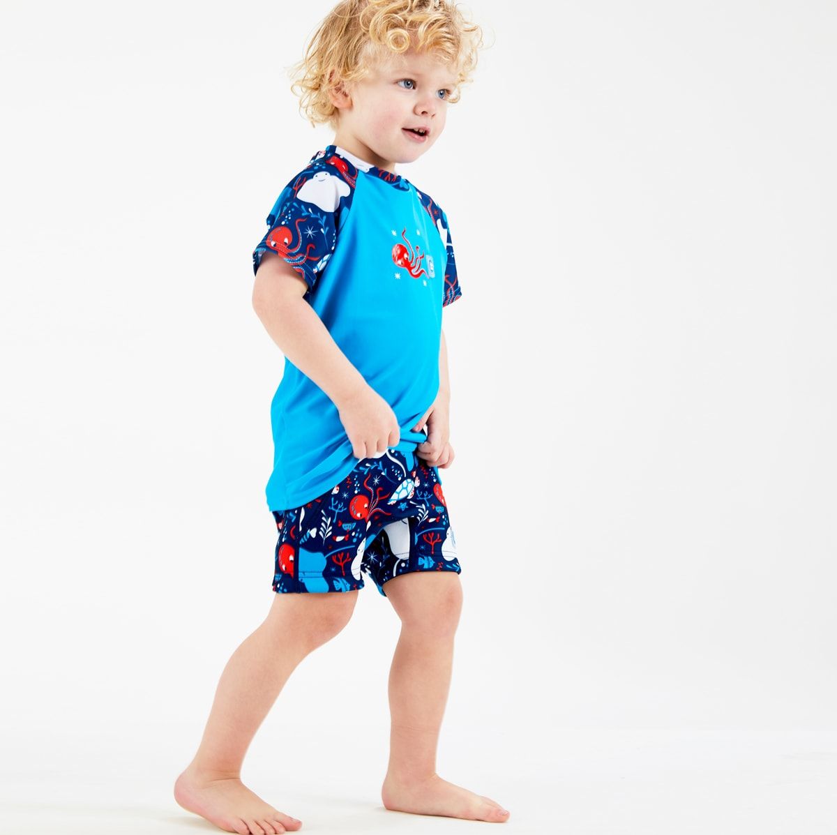 Lifestyle image of toddler wearing UV protective short sleeve rash top in blue, and under the sea themed print on the chest and sleeves. He's also wearing matching jammers or swim shorts.