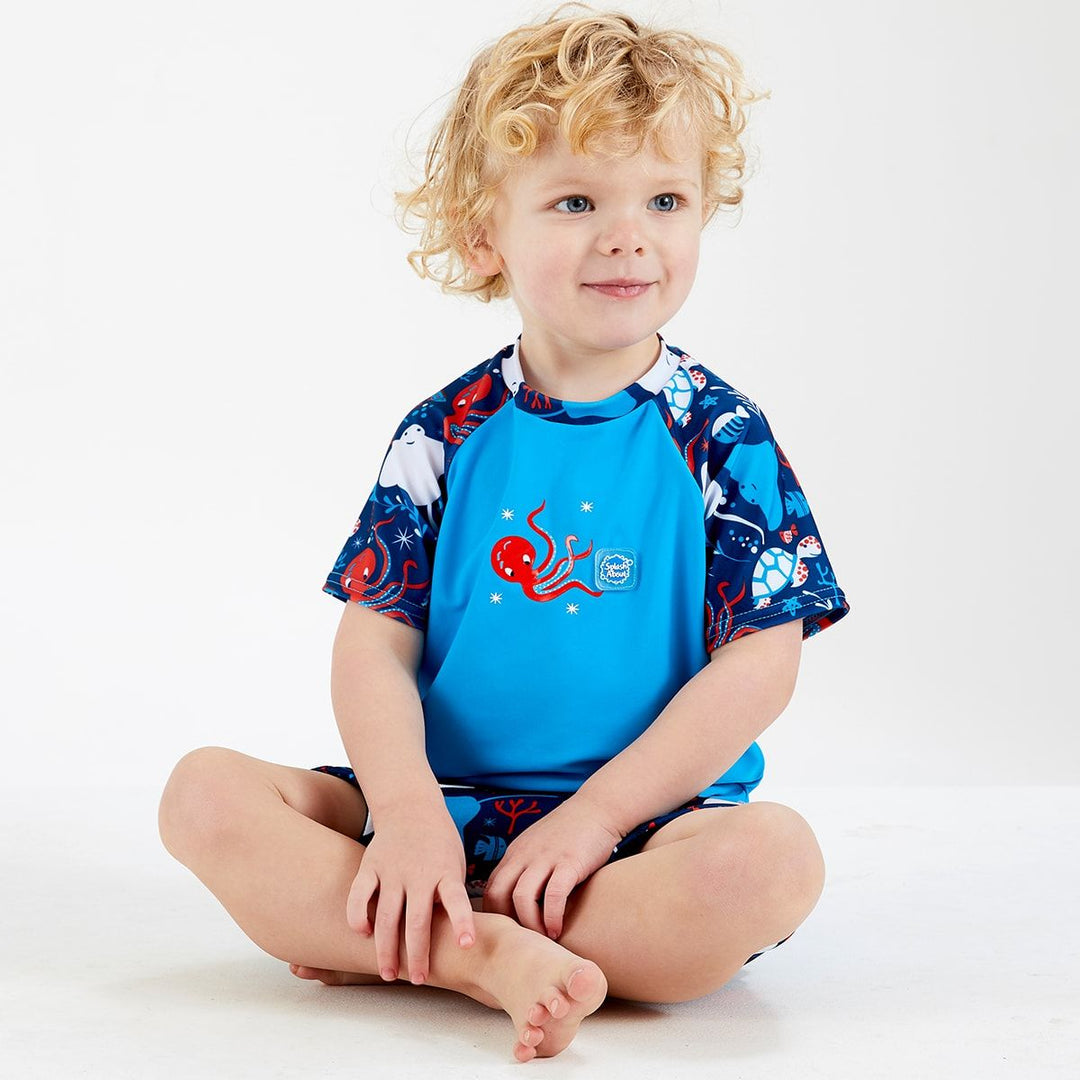 Lifestyle image of toddler wearing UV protective short sleeve rash top in blue, and under the sea themed print on the chest and sleeves. He's also wearing matching jammers or swim shorts.